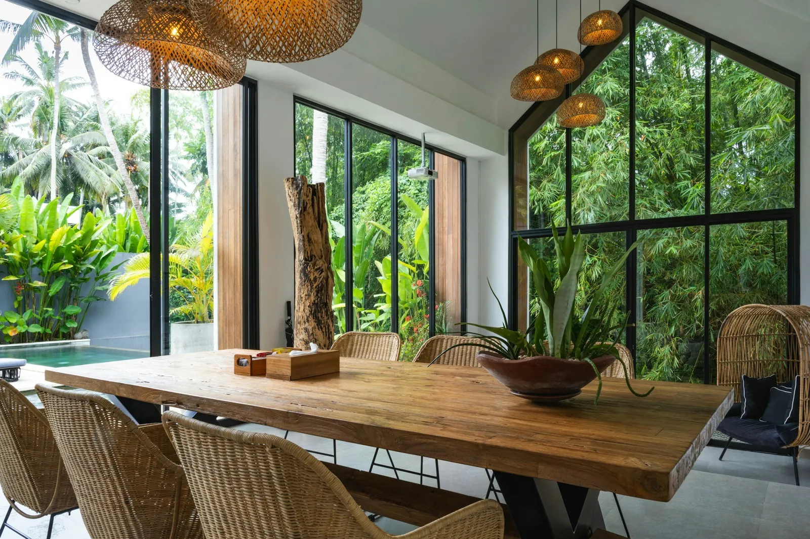 A dining room with a long timber table, woven pendants and full-height glass onto jungle