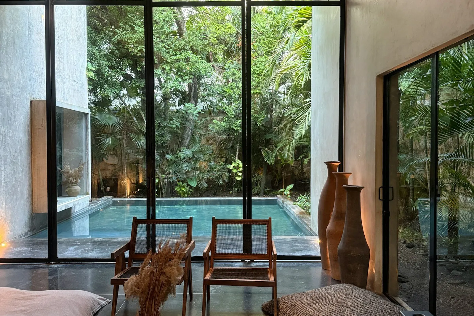 A living room with concrete walls and low seating, a pool and greenery beyond