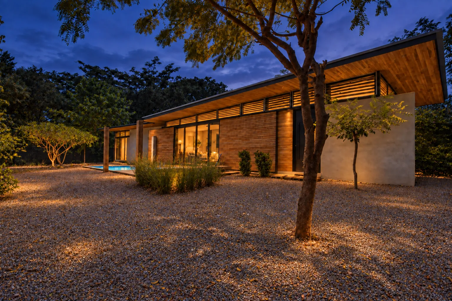 The house at dusk, its timber eave and louvres lit from inside, behind a gravel garden and a tree